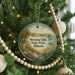 A round ornament hanging on a Christmas tree, featuring a scenic landscape with a path, trees, and a body of water.