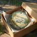 A decorative ornament in a wooden box, featuring a scenic landscape with mountains, trees, and a river.