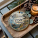 A decorative ornament featuring a scenic landscape of Morro Bay State Park, California, with a candle and pine cones on a wooden tray.