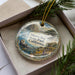 A decorative ornament in a box, featuring a scenic landscape with mountains, water, and wildlife.