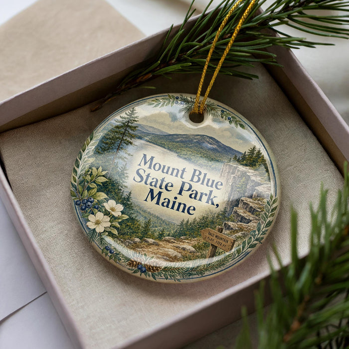 A decorative ornament in a box, featuring a scenic landscape of Mount Blue State Park, Maine.