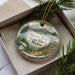 A decorative ornament in a box, featuring a scenic landscape of Mount Blue State Park, Maine.