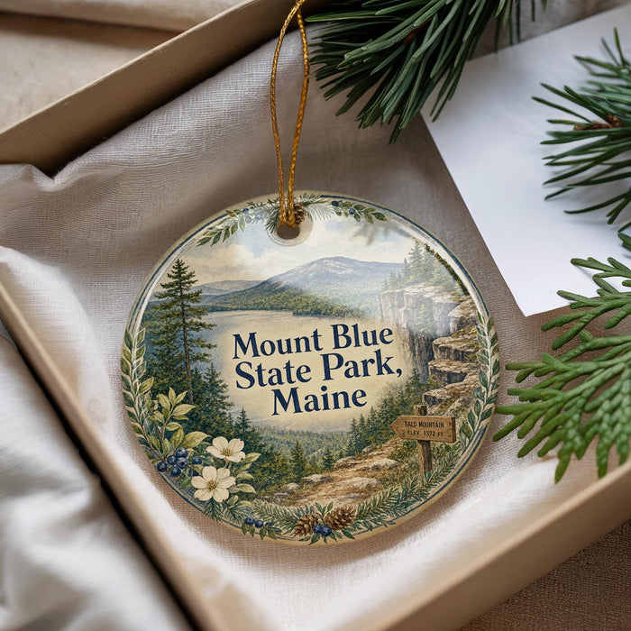 A decorative ornament with a scenic mountain landscape and pine trees, featuring the text "Mount Blue State Park, Maine" and a small sign indicating the location.