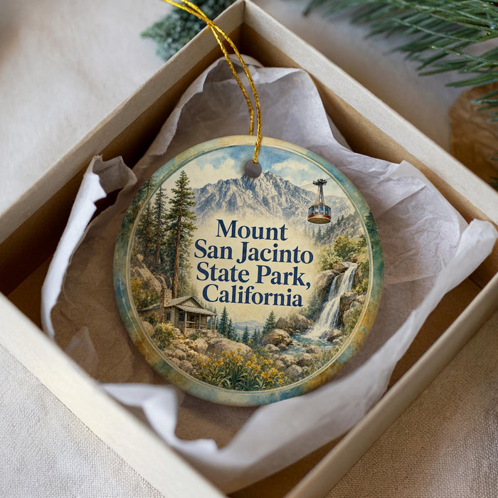 A round ornament with a scenic mountain landscape and a ski lift, placed in a box with a white tissue paper lining.
