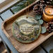 A wooden tray with a decorative ornament featuring a scenic mountain landscape, pine cones, and a lit candle.