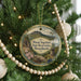 A round ornament hanging on a Christmas tree, featuring a scenic landscape with mountains, pine trees, and a lighthouse.