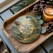 A wooden tray with a decorative ornament featuring a mushroom, rock, and trees, along with pine cones and a lit candle, creating a cozy and rustic atmosphere.