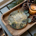 A decorative ornament with a scenic image of Kentucky State Park, featuring a house, trees, and a gate, is displayed on a wooden tray alongside pine cones and a lit candle.