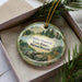 A decorative ornament with a forest scene and a deer, placed in a box with pine branches.