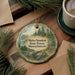 A round ornament with a forest scene and text, placed on a wooden surface next to a cup of coffee and some pine branches.