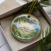 A decorative ornament with a tropical landscape design, featuring a waterfall and lush vegetation, is displayed in a box with pine branches.