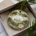 A decorative ornament with a floral design and text, placed in a box with pine branches.