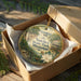 A decorative ornament in a box, featuring a scenic image of a park with a house and a bridge.