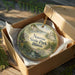 A decorative ornament in a box, featuring a scenic landscape with a bridge and buildings, with the text "Nauvoo State Park, Illinois" printed on it.