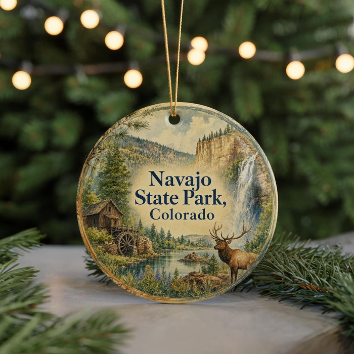 A round ornament with a scenic landscape of Navajo State Park, Colorado, featuring a waterfall, pine trees, and a deer.