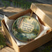 A round ornament in a brown box, featuring a scenic image of the Navarro River and Redwood State Park in California.