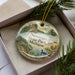 A decorative ornament in a box, featuring a scenic landscape with a lake, trees, and a bird, along with the text "Newaygo State Park, Michigan".