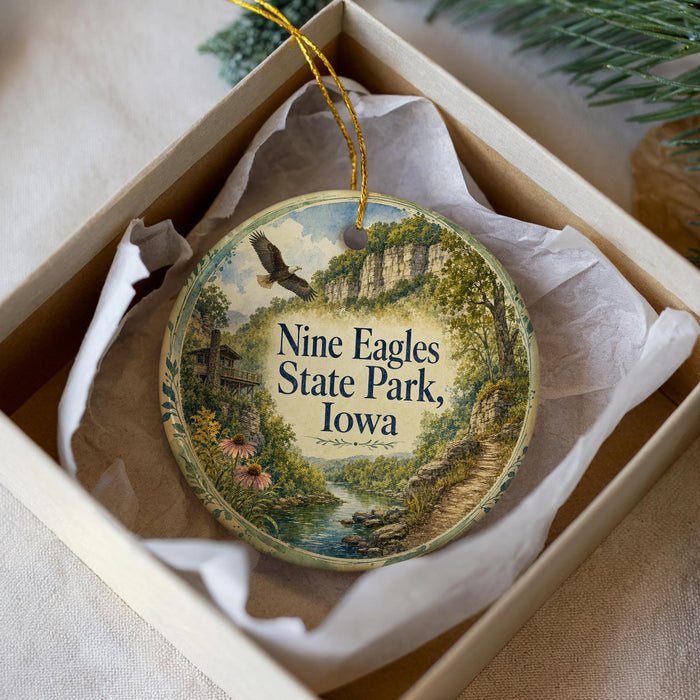 A decorative ornament in a white box, featuring a scenic landscape with a river, trees, and a bird in flight.