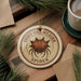 A round ornament with a spider design, featuring the text "SPINY ORB-WEAVER SPIDER" on it, is displayed on a wooden surface alongside a cup of coffee and some pine branches.