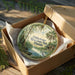 A decorative ornament in a box, featuring a scenic landscape with a river, palm trees, and a bird, is placed on a wooden surface.