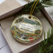A decorative ornament in a box, featuring a scenic landscape of mountains, trees, and a stream.