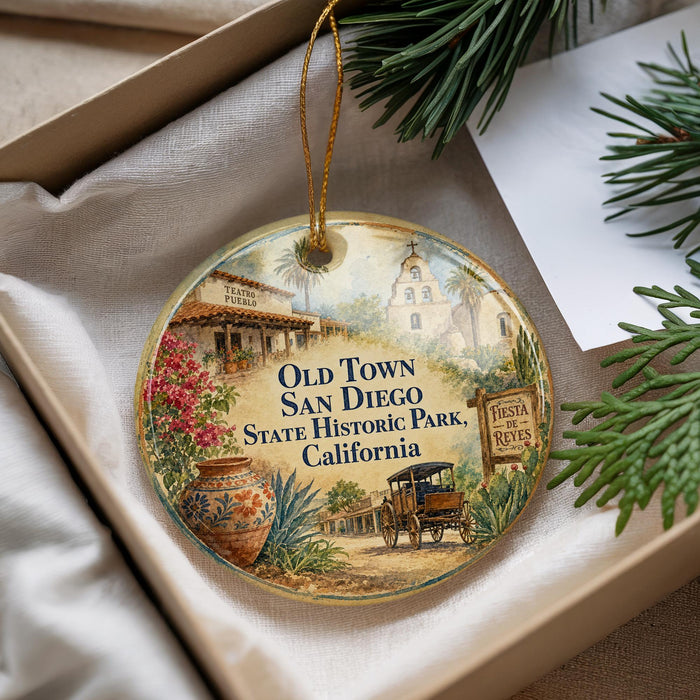 A decorative ornament with a vintage-style illustration of a historical town, featuring a church, a wagon, and other elements, is displayed in a box with pine branches.