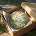 A circular ornament with a bird and a boat, placed in a cardboard box with a gold string.
