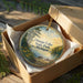 A round ornament in a box, featuring a scenic landscape of Otsego Lake, State Park, and Michigan.