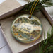 A decorative ornament with a landscape scene featuring a river, mountains, and a building, placed inside a box with pine branches.