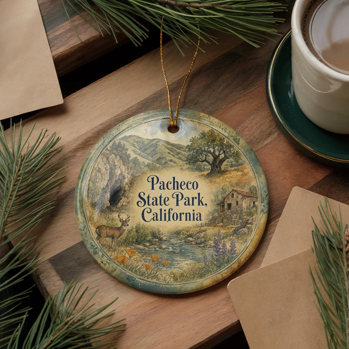 A decorative ornament with a scenic landscape of Pacheco State Park, California, featuring a deer, a river, and mountains. The ornament is hanging from a string and is placed on a wooden surface, surrounded by pine branches and a cup of coffee.