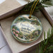 A decorative ornament in a box, featuring a scenic landscape with a lake, trees, and a bird.