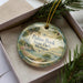 A decorative ornament in a box, featuring a scenic landscape with a lake, mountains, and pine trees.