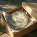 A decorative ornament in a brown box, featuring a scenic landscape with a lake and mountains, is placed on a wooden surface.