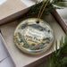 A decorative ornament in a box, featuring a scenic landscape with mountains, pine trees, and flowers, along with the text "Palomar Mountain State Park, California".