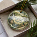 A decorative ornament in a box, featuring a scenic landscape with a waterfall, a bridge, and people walking along a path.