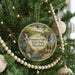 A round ornament hanging on a Christmas tree, featuring a scenic landscape with a waterfall and a bridge.