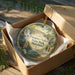 A decorative ornament in a wooden box, featuring a scenic landscape with a waterfall and a bridge, with the text "Patapsco Valley State Park Maryland" printed on it.