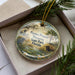 A decorative ornament in a box, featuring a scenic landscape with a bird, palm tree, and bison, along with the text "Paynes Prairie Preserve State Park, Florida".