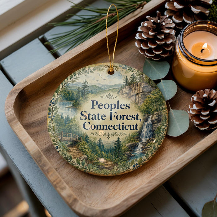 A decorative ornament with a forest scene and text, placed on a wooden tray next to pine cones and a lit candle.