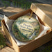 A decorative ornament in a cardboard box, featuring a scenic landscape with a waterfall and forest.