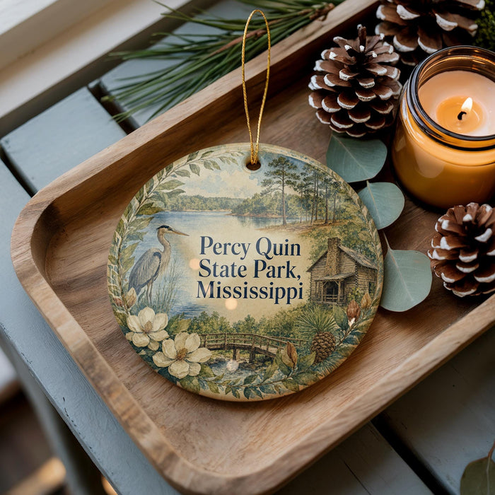A decorative ornament with a scenic image of Perry Quin State Park, Mississippi, featuring a bird, flowers, and a cabin, is displayed on a wooden tray alongside pine cones and a lit candle.