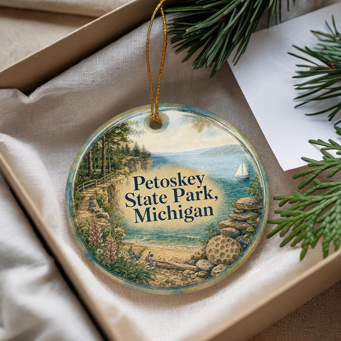 A decorative ornament with a scenic landscape of Petoskey State Park, Michigan, featuring a sailboat on the water and a rocky shoreline.