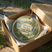 A decorative ornament in a wooden box, featuring a scenic landscape with mountains, trees, and a waterfall.