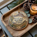 A decorative ornament featuring a desert landscape with a cactus and mountains, along with pine cones and a lit candle, placed on a wooden surface.
