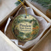 A round ornament with a scenic forest landscape and a bird perched on a sign, placed in a box with a white tissue paper lining.