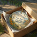 A round ornament in a box, featuring a scenic landscape with mountains, flowers, and a bird, along with the text "Point Mugu State Park, California".
