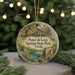 A round ornament with a painting of a house and trees, surrounded by greenery, hangs in front of a blurred background with twinkling lights.
