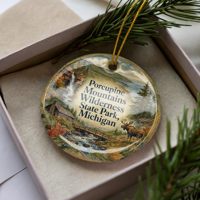 A decorative ornament featuring a scenic landscape with a moose, a waterfall, and a cabin, placed in a box with pine branches.