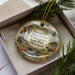 A decorative ornament featuring a scenic landscape with a moose, a waterfall, and a cabin, placed in a box with pine branches.