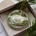 A decorative ornament in a box, featuring an illustration of a forest scene with a bird and a deer.